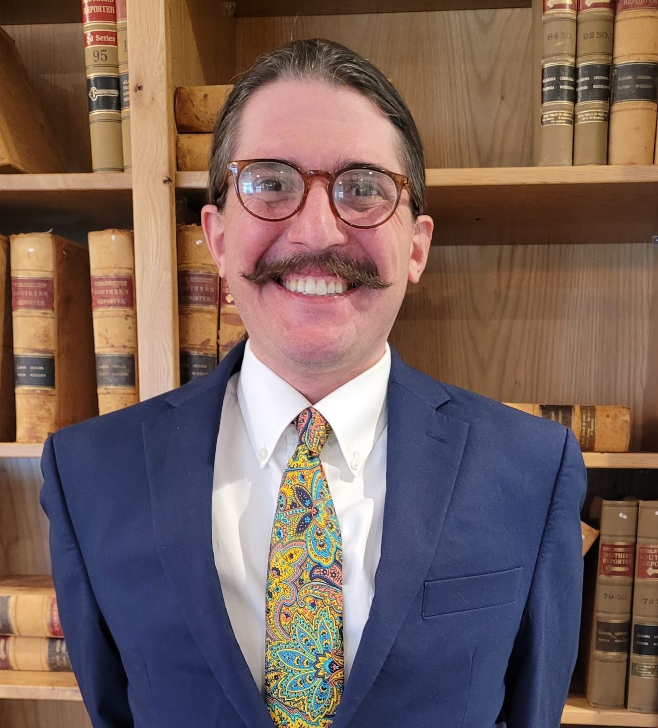 white man with mustache and glasses wearing suit, smiling standing against bookshelf